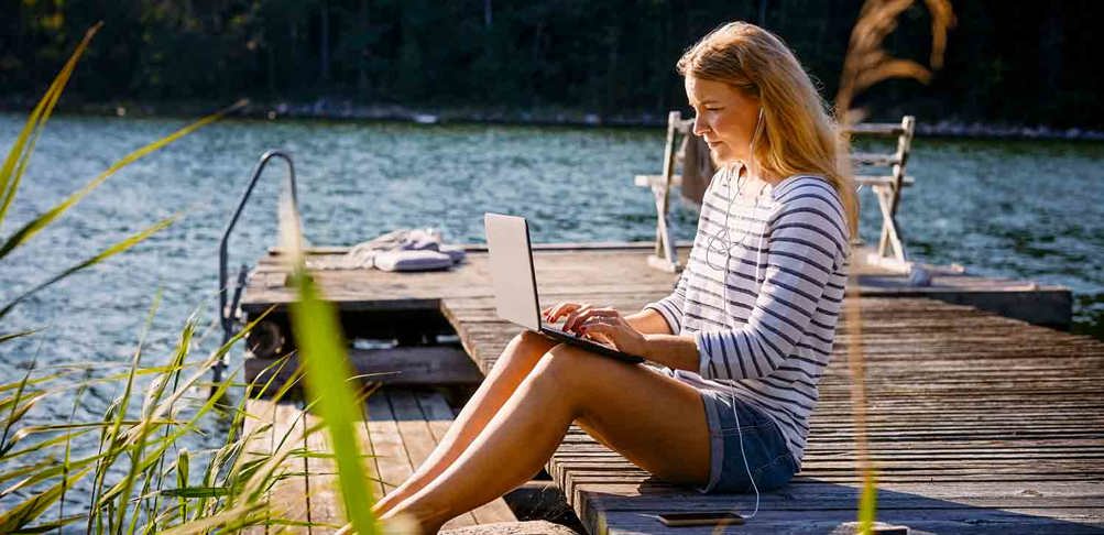 Kvinna med dator sitter på brygga vid vattnet.