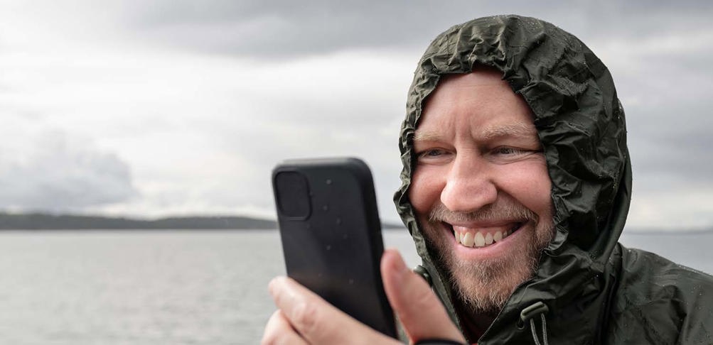 Man som ler när han ser på mobil vid vattnet.