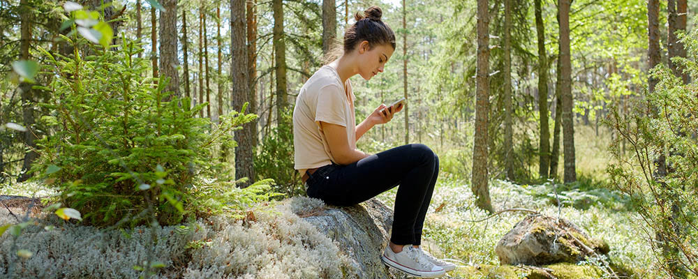 Kvinna med mobiltelefon sitter på en sten i skogen.
