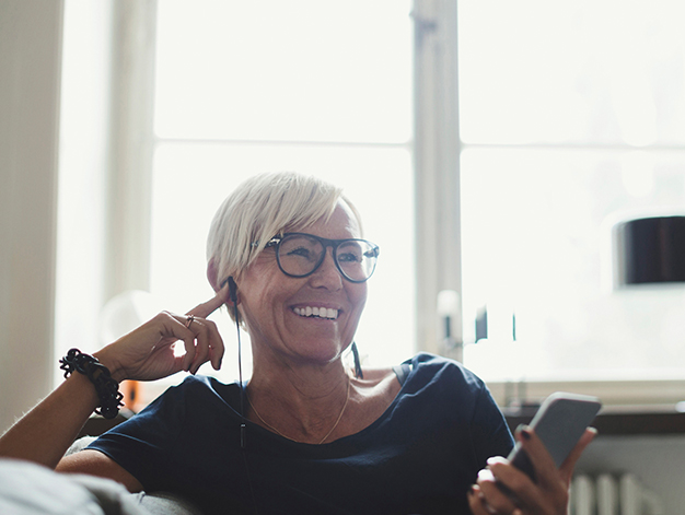 En kvinna sitter i en soffa och ler med mobiltelefon i handen och lyssnar i ett headset