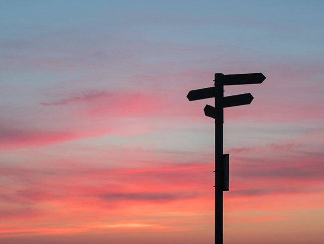 Vägskylt med pilar som pekar åt olika håll i motljus mot en röd himmel.